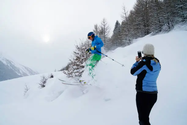 Photo of Young man skis down slope as woman photographs him.