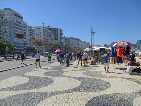 Tourists and locals enjoying the sun on famous Copacabana Beach in Rio de Janeiro. Merchandise in the form of beach towels and other goods is visible.