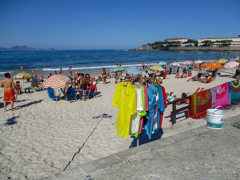 Tourists and locals enjoying the sun on famous Copacabana Beach in Rio de Janeiro. Merchandise in the form of beach towels and other goods is visible.