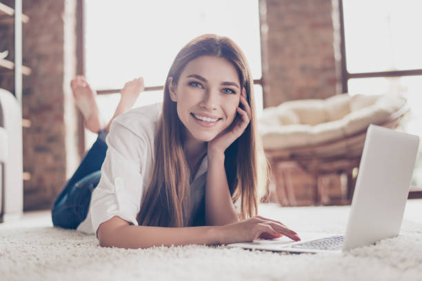 so comfortable and easy! close up of young happy girl typing in her laptop lying on the floor on a white carpet indoors at home - lying down women laptop freedom imagens e fotografias de stock