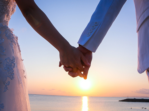 Wedding couple on the sea beach watching sunset. Sunny summer photo. Bride with hair down in off shoulder dress with train. Ocean romantic ceremony. Seaside sunrise love story. Honeymoon.