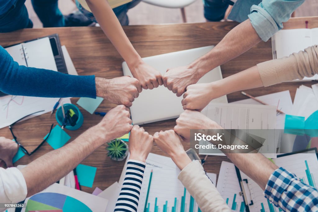 Chiudi la foto ritagliata dei partner che mettono insieme i pugni in un cerchio in cima al tavolo con cose da lavoro. Fiducia, amicizia, unità, concetto di cooperazione - Foto stock royalty-free di Lavoro di squadra