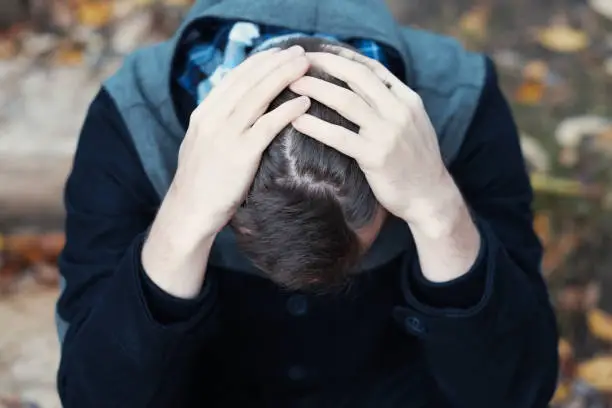 Young man covers his face with his hands in grief or pain. He is sitting outdoor.