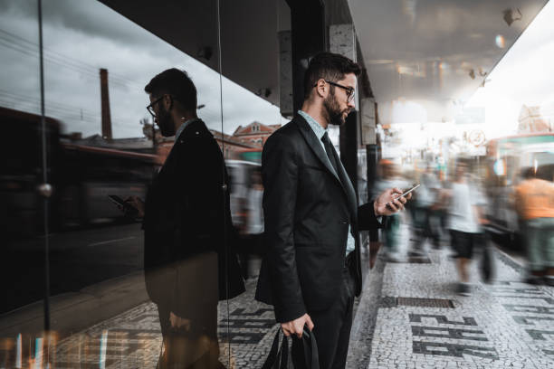 Businessman on street using cellphone Handsome young caucasian bearded man employer in a formal suit is standing next to a bus stop and texting message on the smartphone, surrounded by city fussiness, walking people, and passing buses stirring stock pictures, royalty-free photos & images