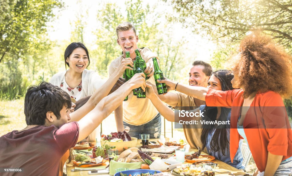 Young multiracial friends toasting at barbecue garden party - Friendship concept with happy people having fun at backyard bbq summer camp - Food and drinks fancy picnic lunch - Focus on beer bottles Beer - Alcohol Stock Photo