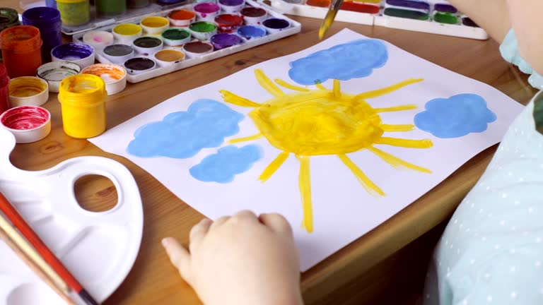 Cute young girl draws yellow sun and blue clouds. Painting concept