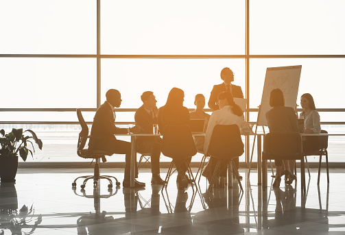 Group of people sitting at the office table while their partner standing near board with chart. Panoramic window on background