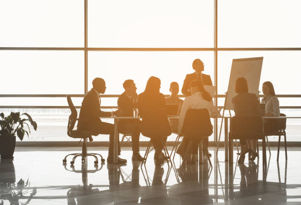 Man making startup analysis to colleagues Group of people sitting at the office table while their partner standing near board with chart. Panoramic window on background plank stock pictures, royalty-free photos & images