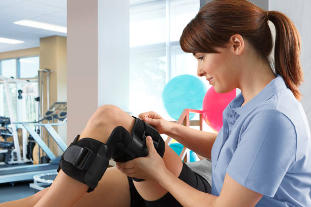 Female Physical Therapist Adjusting Knee Brace On Female Patient A female physical therapist in her mid thirties adjusts the knee brace of a patient who is lying on a treatment table in a clinical setting. knee brace stock pictures, royalty-free photos & images