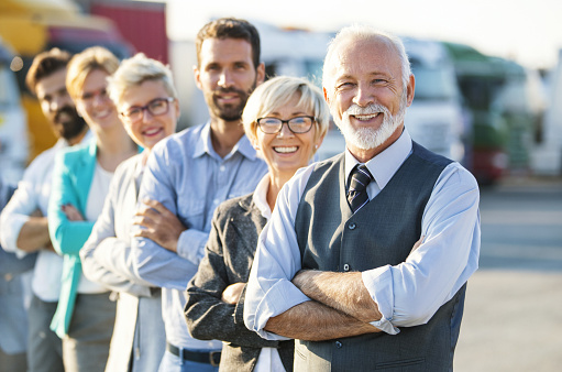 Closeup front view of multi aged corporate team standing in a row in front of their company. There are many semi trucks in background. Senior executive is in front, two more men and three women behind him.