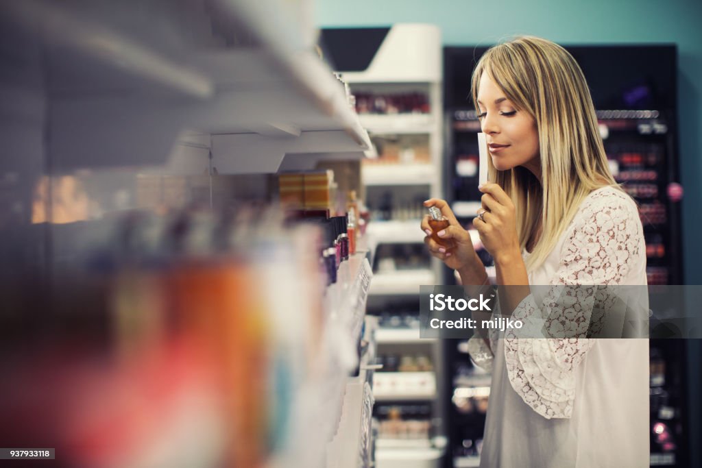 Young woman shopping at cosmetics and make-up shop Beautiful young woman choosing and testing perfume in cosmetics retail shop. She is beautiful, fresh and looks very happy while shopping Perfume Stock Photo