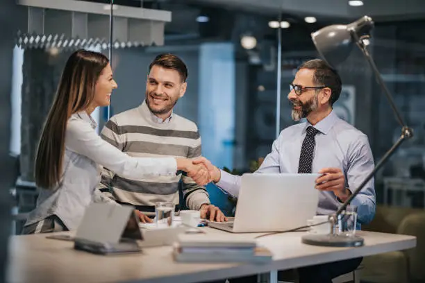 Happy couple having a meeting with their real estate agent while woman and mature man are shaking hands. Focus is on men.