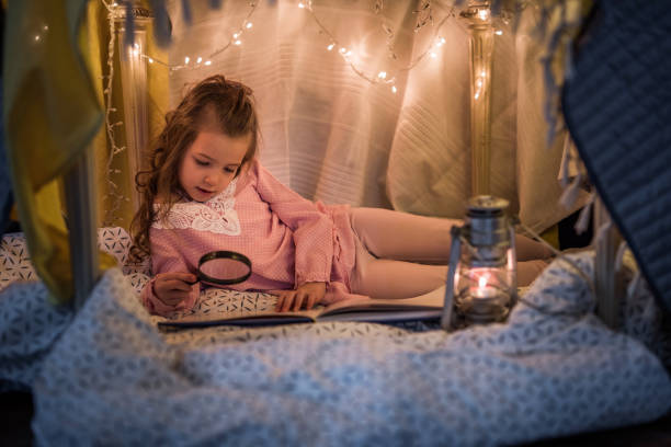 istock Small girl using magnifying glass while reading a book in a tent. 938027072