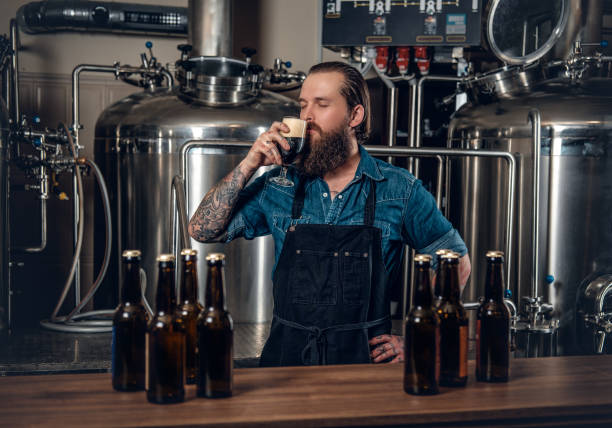 A man manufacturer tasting beer in the microbrewery. Portrait of tattooed, bearded hipster male manufacturer tasting beer in the microbrewery. microbrewery stock pictures, royalty-free photos & images