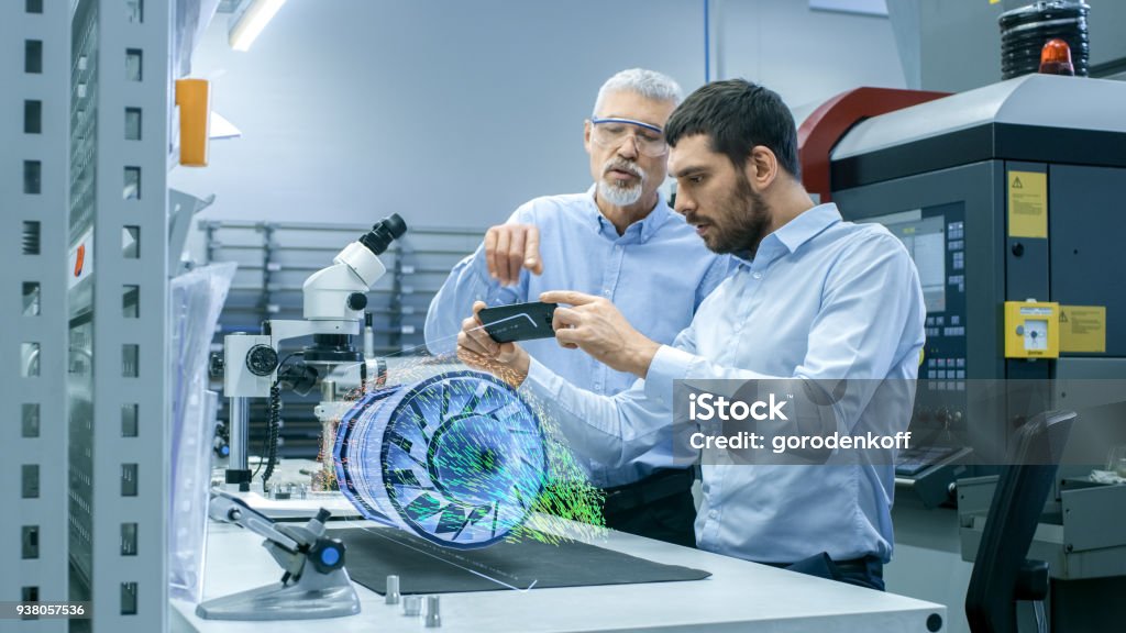 Two Engineers Works with Mobile Phone Using Augmented Reality Holographic Projection 3D Model of the Engine Turbine Prototype. Development of Virtual Mixed Reality Application. Augmented Reality Stock Photo Two Engineers Works with Mobile Phone Using Augmented Reality Holographic Projection 3D Model of the Engine Turbine Prototype. Development of Virtual Mixed Reality Application. Augmented Reality Stock Photo