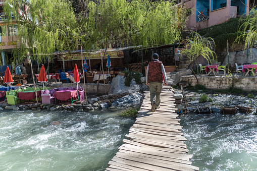 A Berber man walks over an old wooden bridge over fast flowing stream in a valley in the High Atlas Mountains, Morocco. Cafe seating on the banks of the river.