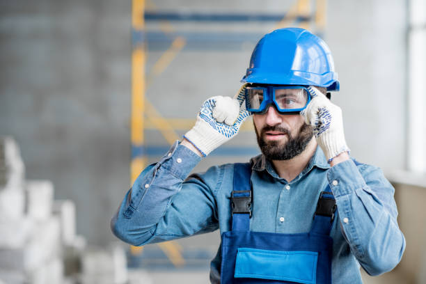 Builder in uniform indoors Close-up portrait of a handsome bearded builder with protective glasses and helmet indoors helmet hardhat protective glove safety stock pictures, royalty-free photos & images