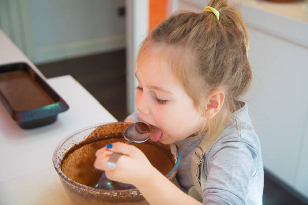 four years old girl tasting whipped chocolate cream from glass bowl with spoon, ready to cook a sponge chocolate cake at kitchen home