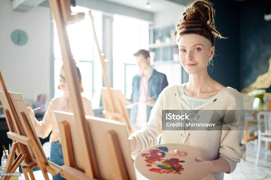 Student painting Happy young student with palette looking at camera while painting on easel in studio of arts Artist Stock Photo