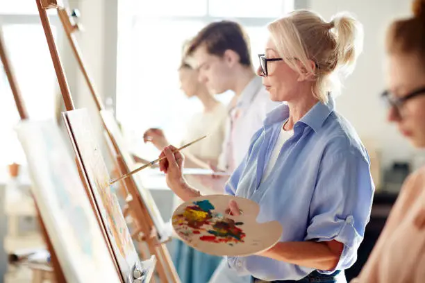 Blonde mature woman painting picture on easel with mixed oil colors between her students