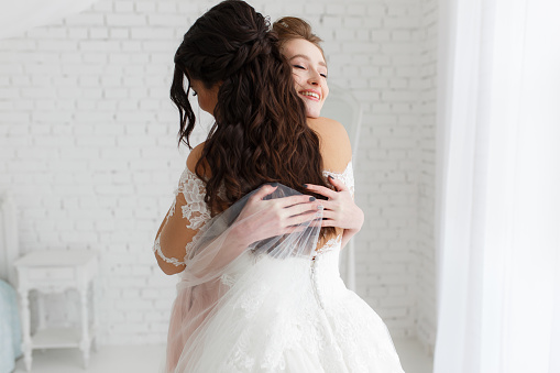 luxury bride hugging bridesmaid and smiling, joyful moment in minimalistic loft white brick background.