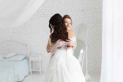 luxury bride hugging bridesmaid and smiling, joyful moment in minimalistic loft white brick background.