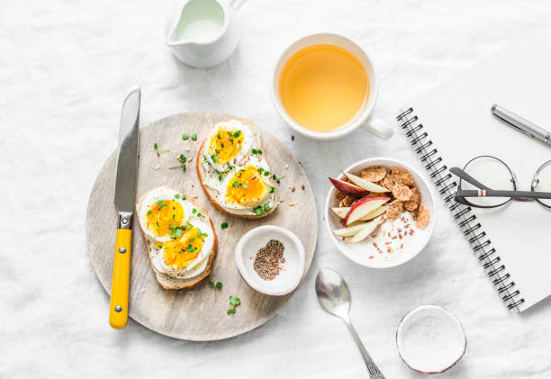 istock Morning breakfast table inspiration - sandwiches with cream cheese and boiled egg, yogurt with apple and flax seeds, herbal detox tea, notebook, glasses on light background, top view. Flat lay 938158976