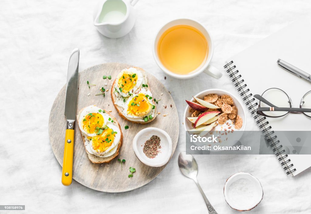 Morning breakfast table inspiration - sandwiches with cream cheese and boiled egg, yogurt with apple and flax seeds, herbal detox tea, notebook, glasses on light background, top view. Flat lay Breakfast Stock Photo