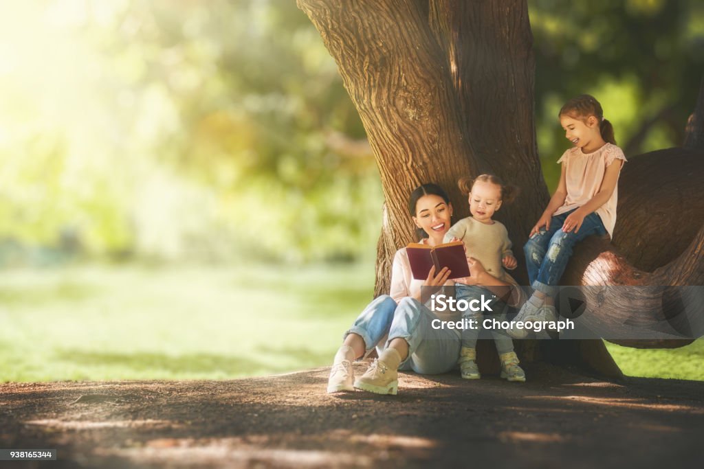mom reading a book to her children Mother and daughters sitting under the tree on summer lawn. Happy family playing outdoors. Pretty young mom reading a book to her children in the park outside. Reading Stock Photo