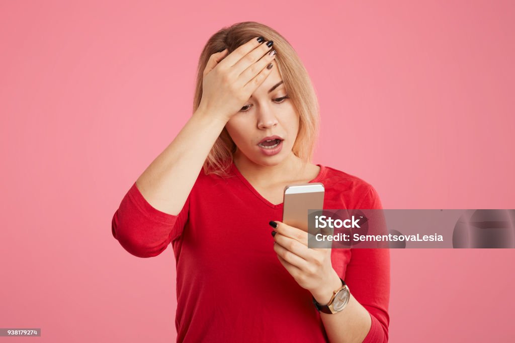 Terrfied blonde young woman stares at camera of mobile phone, keeps hand on forehead, reads bad news on internet website, isolated over pink background. People, emotions and technology concept Adult Stock Photo