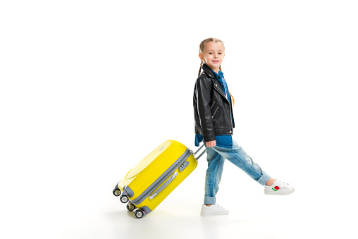 Side view of little tourist with pigtails carriyng her yellow wheel suitcase isolated on white