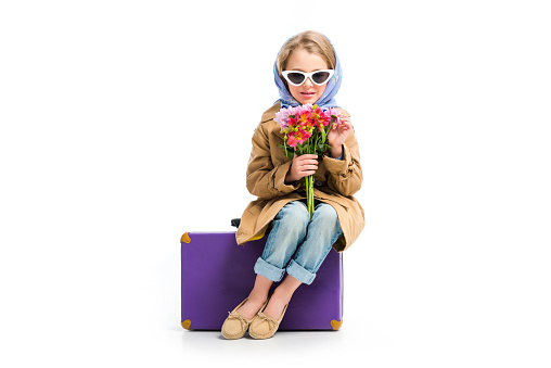 Stylish child in sunglasses and headscarf holding flowers while sitting on suitcase isolated on white