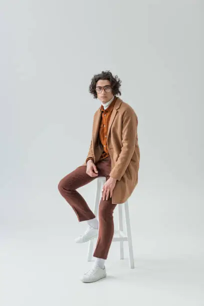 Photo of Young stylish man in eyeglasses sitting on chair isolated on grey
