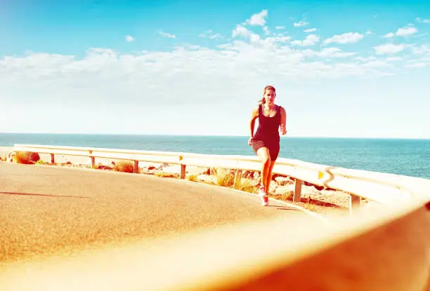 Sporty female runs on a road near the sea in a tropical climate while the sun shines. Her exercising is pushing her endurance for marathon running or a triathlon. She looks calm, as she runs in the heat.