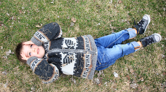 little boy lies on grass, with sweater covered in dry grass, gazing up at the clouds