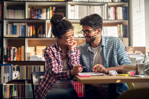 istock Flirty attractive beautiful hardworking stylish hipster young student couple reading a book together while sitting in the library or classroom near the sunny window. 938307894