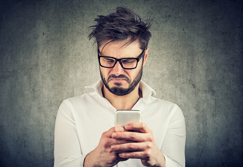 Young man in eyeglasses surfing social media and looking confused and angry watching news.