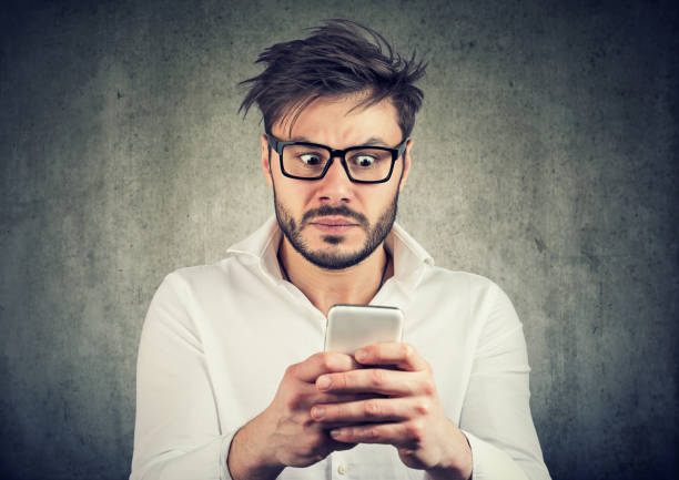 Scared man watching social media Young bearded man in glasses watching smarpthone and looking scared with bad news. slander stock pictures, royalty-free photos & images