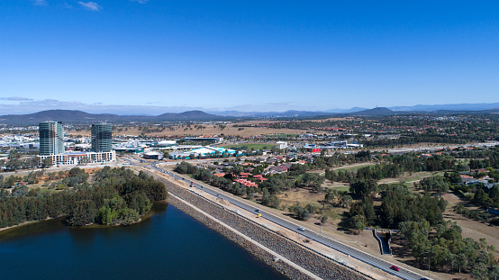 Aerial drone photograph, high view point from Yerrabi Pond in foreground towards Gungahlin town centre, a few people walking around, cars moving and parked, homes, buildings with recognisable commercial properties plus football stadium in view on a nice sunny day.