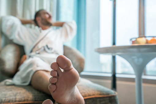 Cheerful young man relaxing on sofa, wearing bathrobe in hotel room watching the city view through window. City of Kuala Lumpur, Malaysia, travel Asia concept