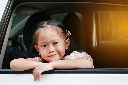 Cute little asian girl looking camera from the window of the car with rays of sunlight.