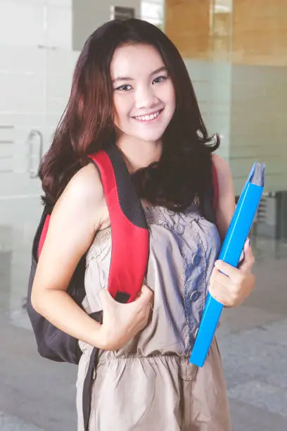 Female college student smiling at the camera while carrying a folder and standing in the school