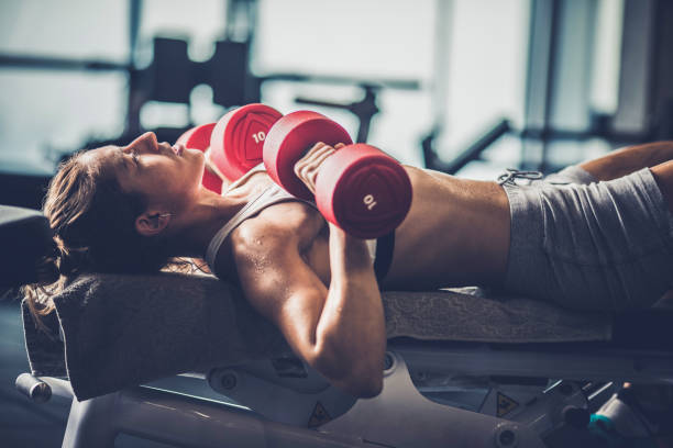 Athletic woman having a weight training on a bench in a gym. Female athlete lying on a bench and having chest exercises with dumbbells in a gym. chest dumbbells ripl fitness
