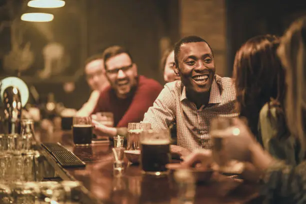 Group of people drinking beer at a bar counter. Focus is on happy black man talking to his friend.