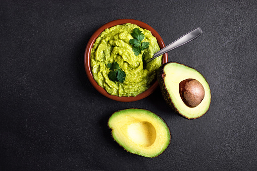 Guacamole sauce  in clay bowl with  cut half avocado on dark background. Top view with copy space