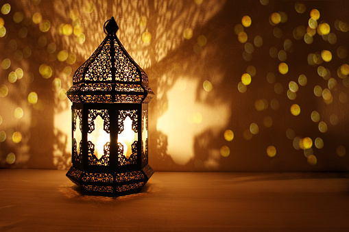 Ornamental Arabic lantern with burning candle glowing at night and glittering golden bokeh lights. Festive greeting card, invitation for Muslim holy month Ramadan Kareem, dark background.