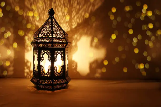 Photo of Ornamental Arabic lantern with burning candle glowing at night and glittering golden bokeh lights. Festive greeting card, invitation for Muslim holy month Ramadan Kareem. Dark background