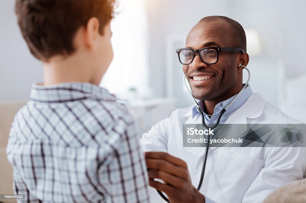 Afro american male doctor examining boys respiratory system Stethoscope exam. Attractive cheerful male doctor listening to boy while putting on glasses and using stethoscope Doctor Stock Photo
