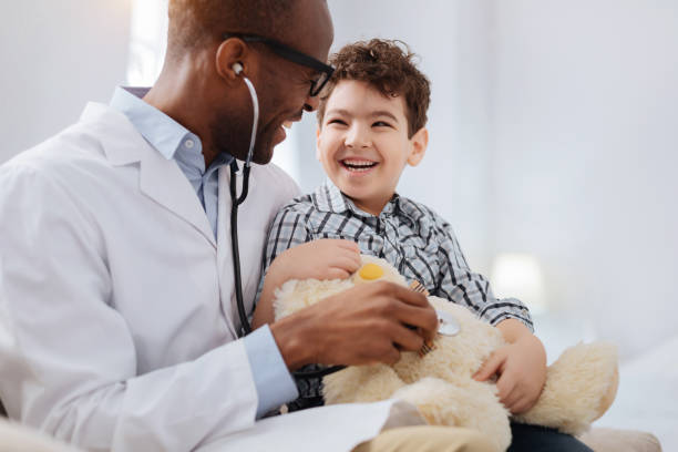 Afro american male doctor diagnosing plush bear Immediate treatment. Enthusiastic vigorous male doctor sitting with boy while listening to plush bear and talking pediatrician stock pictures, royalty-free photos & images