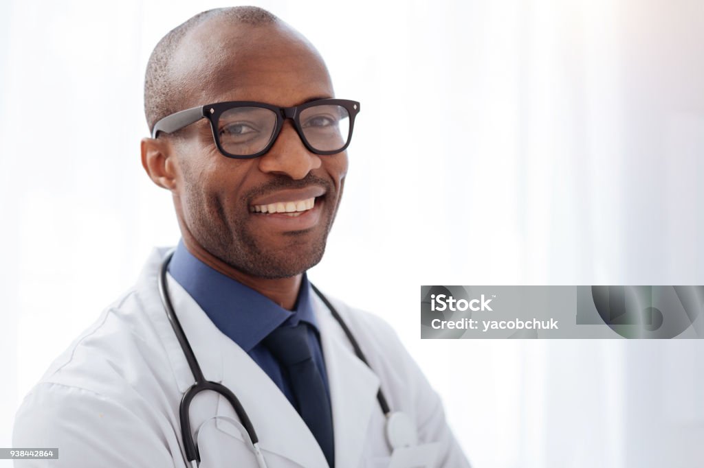 Jovial male doctor smiling to camera Help people. Gay afro american male doctor posing on light background while smiling and gazing at camera Doctor Stock Photo